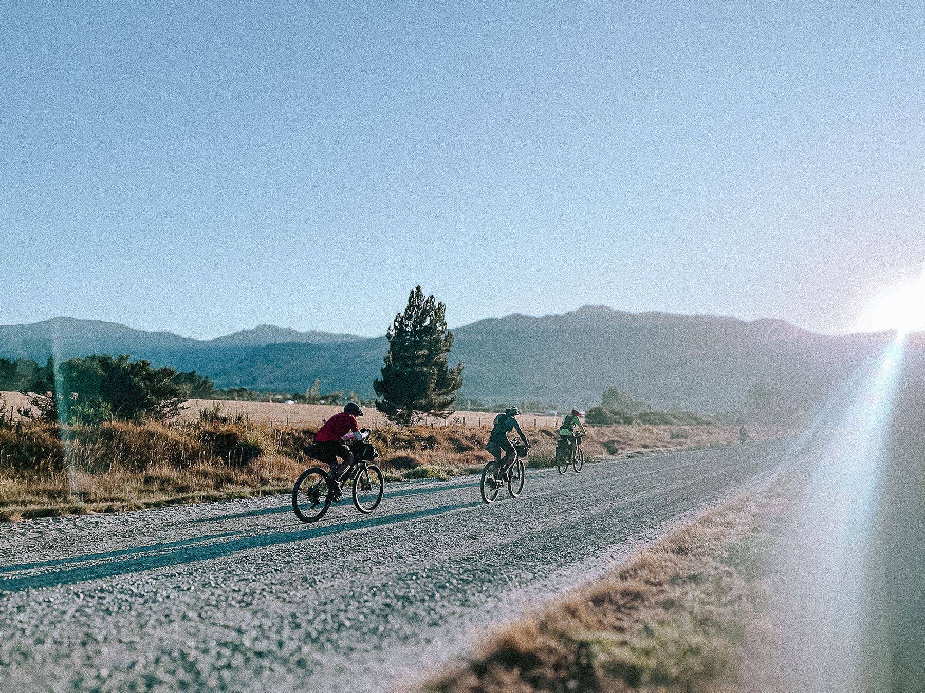 Three bikepackers riding a gravel road at first light, hills behind